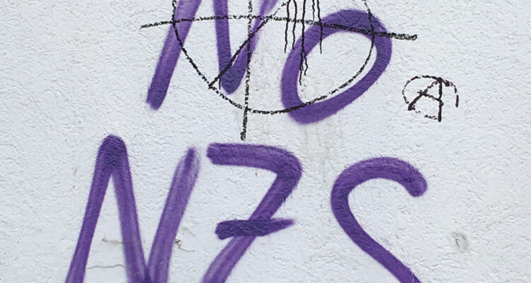 Photo of a graffiti in purple spray-paint on a white wall that spells 'No Nzs', overwritten by a far-right symbol done in feeble black marker