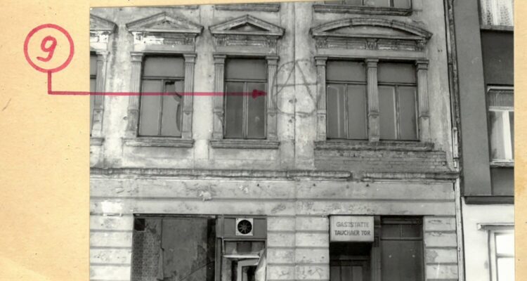 Black-and-white photograph of a façade bearing a spray-painted anarchist symbol between two windows.